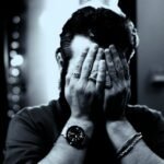 A person covers their face with hands, wearing a watch
